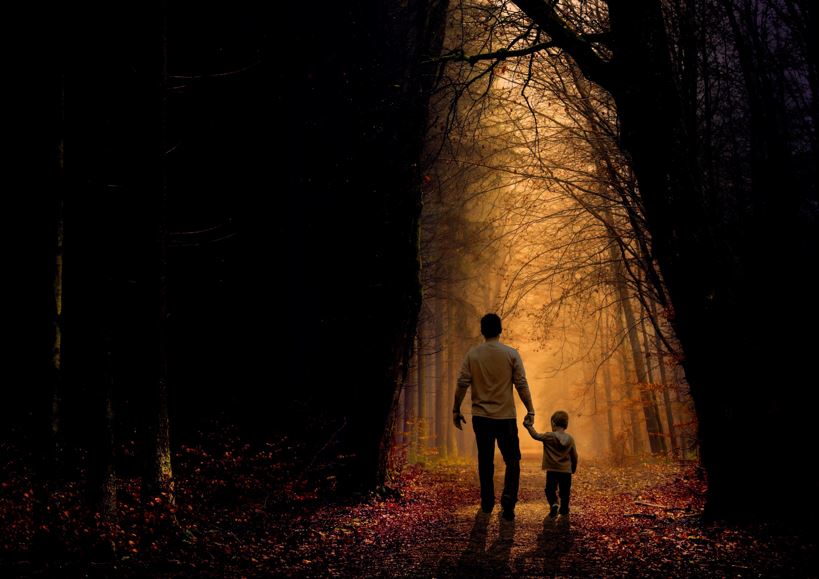 Mies ja lapsi kävelee synkässä metsässä, jossa näkyy myös valoa..